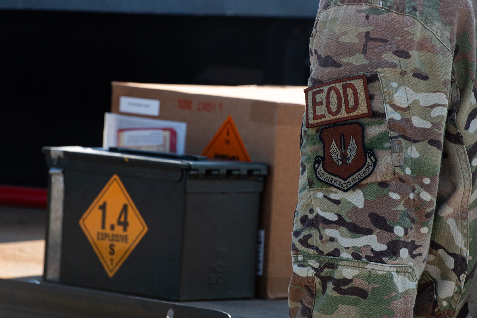 A U.S. Air Force explosive ordnance disposal team member stands in front of an ammunition can displayed on an elevated platform.