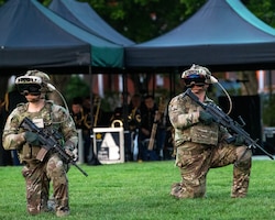 Two Army soldiers in Army Combat Uniforms (green camouflage) are kneeling on one knee on a green lawn. They each have a black rifle in their hands and large helmets on their heads, as well as a large, black goggle devise over their eyes.