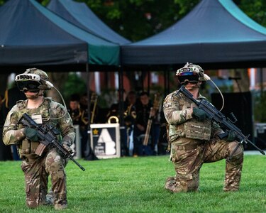 Two Army soldiers in Army Combat Uniforms (green camouflage) are kneeling on one knee on a green lawn. They each have a black rifle in their hands and large helmets on their heads, as well as a large, black goggle devise over their eyes.