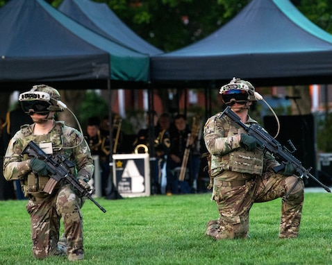 Two Army soldiers in Army Combat Uniforms (green camouflage) are kneeling on one knee on a green lawn. They each have a black rifle in their hands and large helmets on their heads, as well as a large, black goggle devise over their eyes.