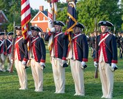Soldiers dressed in Revolutionary War-era uniforms with dark blue coats trimmed in red and white pants and tri-cornered hats are holding the US Flag and US Army flag while standing in a straight line.