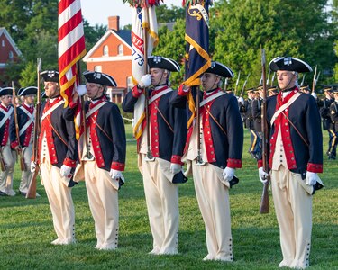 Soldiers dressed in Revolutionary War-era uniforms with dark blue coats trimmed in red and white pants and tri-cornered hats are holding the US Flag and US Army flag while standing in a straight line.