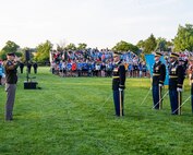 An Army general officer in the Army Green Service Uniform (dark green jacket and khaki colored pants) is standing on a green lawn while saluting toward Army Soldiers on the right side of the picture. The other soldiers are dressed in dark service uniforms, and they are holding swords down in front of them. There is an audience standing on bleachers in the far background.