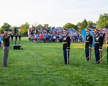 An Army general officer in the Army Green Service Uniform (dark green jacket and khaki colored pants) is standing on a green lawn while saluting toward Army Soldiers on the right side of the picture. The other soldiers are dressed in dark service uniforms, and they are holding swords down in front of them. There is an audience standing on bleachers in the far background.