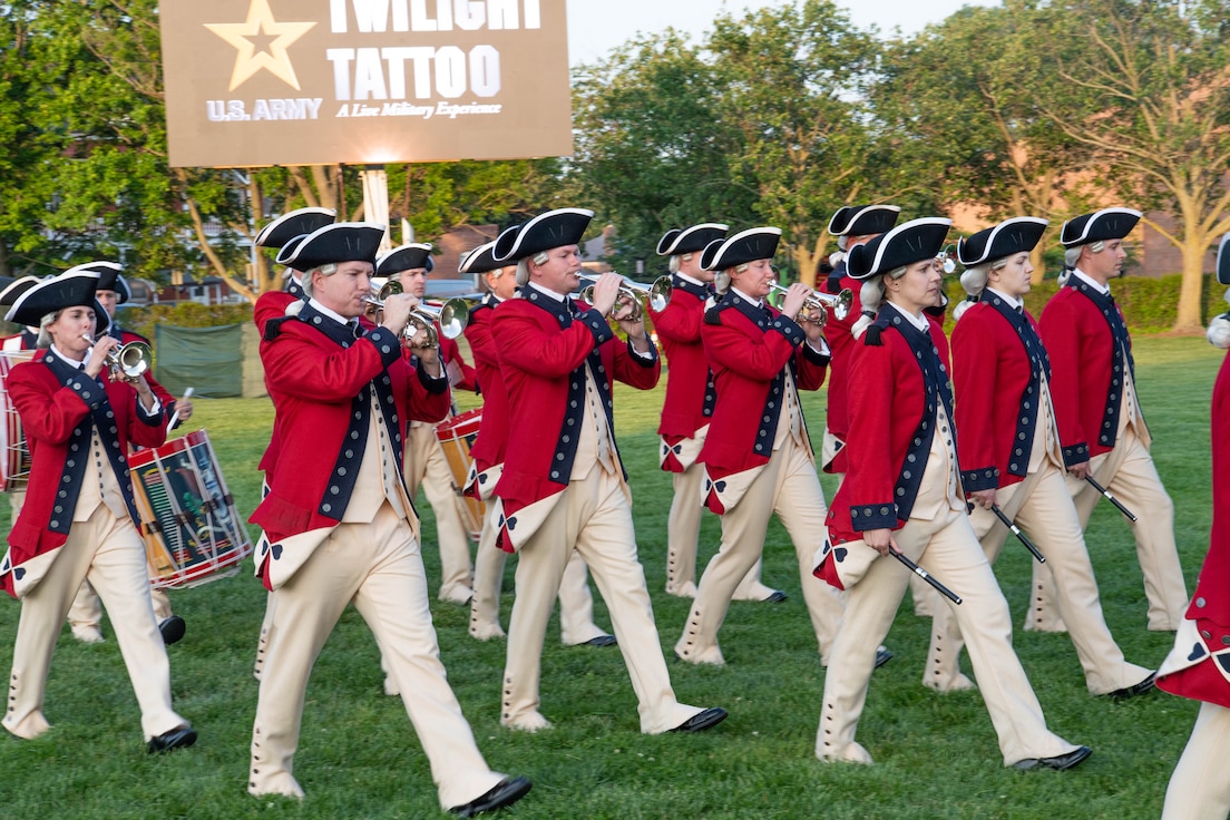 Alt Text
Army soldiers dressed in Revolutionary War-era red with blue trim coats, white pants and tri-cornered hats are marching while playing fifes, bugles and drums. They are on a green lawn with a large screen in the background that is projecting a graphic with the words US Army and Twilight Tattoo.