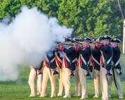 About a dozen Soldiers dressed in Revolutionary War-era uniforms with dark blue coats trimmed in red and white pants and tri-cornered hats are shooting rifles. The smoke from the rifles is billowing in front of them.