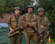 Three Black soldiers in World War 1-era uniforms are standing in a triangle formation. One is carrying a saxophone, another is carrying a trumpet, and the one in front is carrying a rifle.