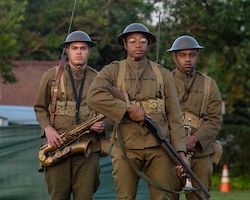 Three Black soldiers in World War 1-era uniforms are standing in a triangle formation. One is carrying a saxophone, another is carrying a trumpet, and the one in front is carrying a rifle.