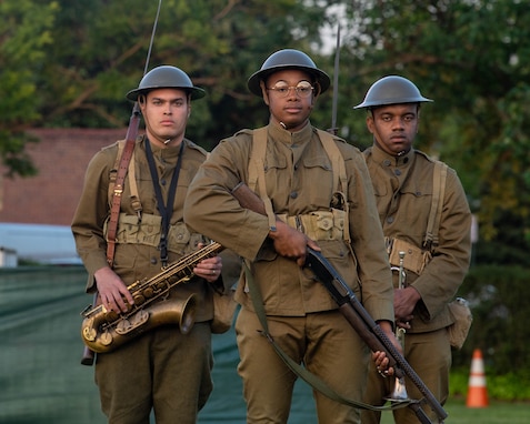 Three Black soldiers in World War 1-era uniforms are standing in a triangle formation. One is carrying a saxophone, another is carrying a trumpet, and the one in front is carrying a rifle.