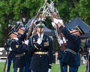 Two rows of Soldiers dressed in dark dress uniforms are extending their bayonet-tipped rifles up toward each other, crisscrossing the tips, as another Soldier carrying a sword pointing upward in front of his face is in the foreground.