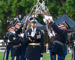 Two rows of Soldiers dressed in dark dress uniforms are extending their bayonet-tipped rifles up toward each other, crisscrossing the tips, as another Soldier carrying a sword pointing upward in front of his face is in the foreground.