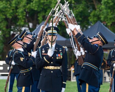 Two rows of Soldiers dressed in dark dress uniforms are extending their bayonet-tipped rifles up toward each other, crisscrossing the tips, as another Soldier carrying a sword pointing upward in front of his face is in the foreground.