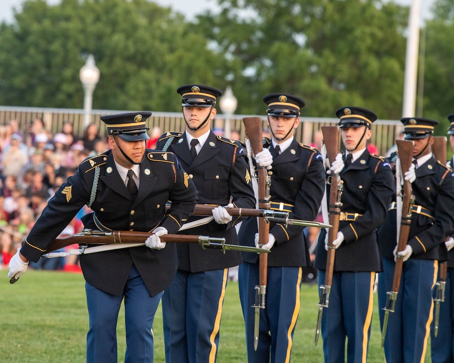 Army Soldiers in dark ceremonial uniforms are standing in a line doing various movements with their rifles.