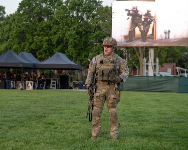 A Soldier dressed in the Army Combat Uniform (green camouflage) with helmet and rifle in hand is performing as an actor at a ceremony on a green lawn. There is a large screen behind him that is projecting his image.