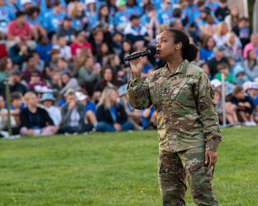 Soldier in Army Combat Uniform (green camouflage) is holding a microphone and singing while standing on a green lawn with an audience in the background sitting on metal bleachers.