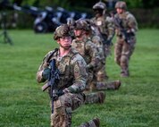 Army soldiers in Army Combat Uniforms (green fatigues) are kneeling on one knee in a long straight row on a green lawn. Some in the background are standing. They each have a black rifle in their hands and helmets on their heads.