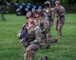 Army soldiers in Army Combat Uniforms (green fatigues) are kneeling on one knee in a long straight row on a green lawn. Some in the background are standing. They each have a black rifle in their hands and helmets on their heads.