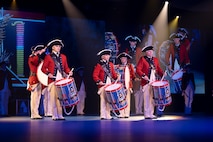 Army soldiers dressed in Revolutionary War-era red with blue trim coats, white pants and tri-cornered hats standing in a group while playing on large drums that they are carrying in front of them using a shoulder strap.