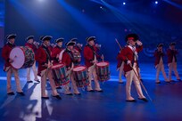Army soldiers dressed in Revolutionary War-era red with blue trim coats, white pants and tri-cornered hats are marching while playing fifes, bugles and drums. The member in front is holding a long stick while saluting and pointing his left foot out.