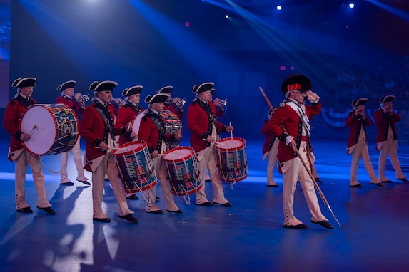 Army soldiers dressed in Revolutionary War-era red with blue trim coats, white pants and tri-cornered hats are marching while playing fifes, bugles and drums. The member in front is holding a long stick while saluting and pointing his left foot out.