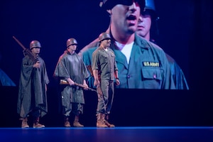 Three Army soldiers dressed in Korean War-era dark green uniforms are standing on a stand speaking during a ceremony. There is a large screen behind them projecting their images.