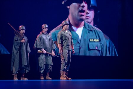 Three Army soldiers dressed in Korean War-era dark green uniforms are standing on a stand speaking during a ceremony. There is a large screen behind them projecting their images.