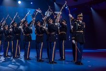 Army Soldiers in dark ceremonial uniforms are standing in two parallel rows with ceremonial rifles raised and tips of rifles crossing each other while one member is standing in the foreground with a sword held by his side.