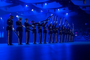 Army Soldiers in dark ceremonial uniforms standing in a straight line are twirling ceremonial rifles.