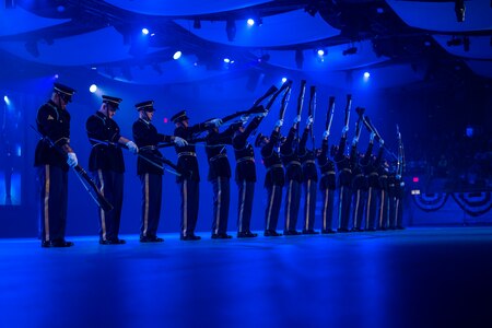 Army Soldiers in dark ceremonial uniforms standing in a straight line are twirling ceremonial rifles.