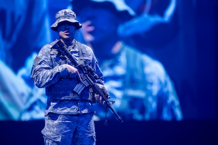 An Army Soldier dressed in the Army Combat Uniform (green camouflage) is standing with a rifle in his hands while speaking a monologue. Behind him is a large screen that is projecting his image.