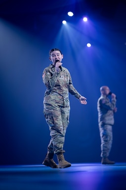Two Soldiers in Army Combat Uniforms are holding microphones and singing on a stage during a ceremony.