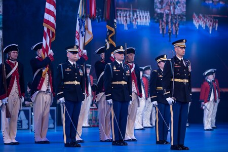 Service members in Army ceremonial uniforms and others wearing blue and red Revolutionary War-era uniforms are standing at attention during a ceremony. The Soldiers in front are holding swords down at their sides. Others in the background are carrying the US and Army flags