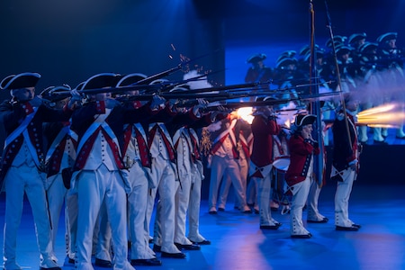 About a dozen Soldiers dressed in Revolutionary War-era uniforms with dark blue coats trimmed in red and white pants and tri-cornered hats are shooting rifles. The smoke and fire from the rifles is billowing in front of them.