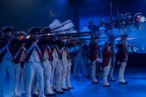 About a dozen Soldiers dressed in Revolutionary War-era uniforms with dark blue coats trimmed in red and white pants and tri-cornered hats are shooting rifles. The smoke from the rifles is billowing in front of them.