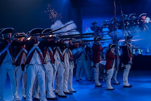 About a dozen Soldiers dressed in Revolutionary War-era uniforms with dark blue coats trimmed in red and white pants and tri-cornered hats are shooting rifles. The smoke from the rifles is billowing in front of them.