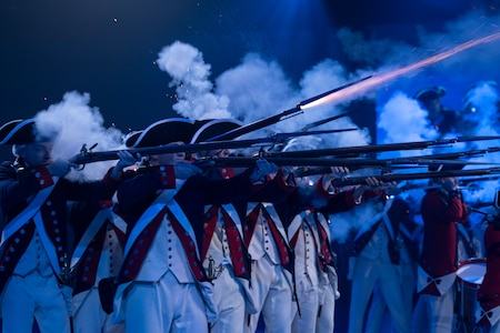 About a dozen Soldiers dressed in Revolutionary War-era uniforms with dark blue coats trimmed in red and white pants and tri-cornered hats are shooting rifles. The smoke from the rifles is billowing in front of them.