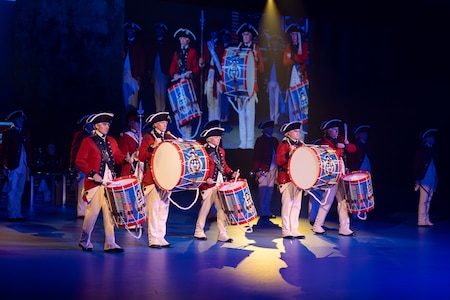 Army soldiers dressed in Revolutionary War-era red with blue trim coats, white pants and tri-cornered hats standing in a group while playing on large drums that they are carrying in front of them using a shoulder strap.