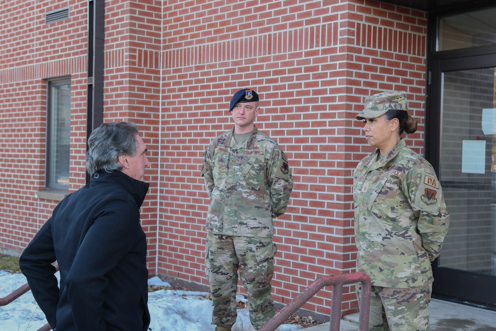 Two military members are greeted by a man in a black coat on a set of stairs.