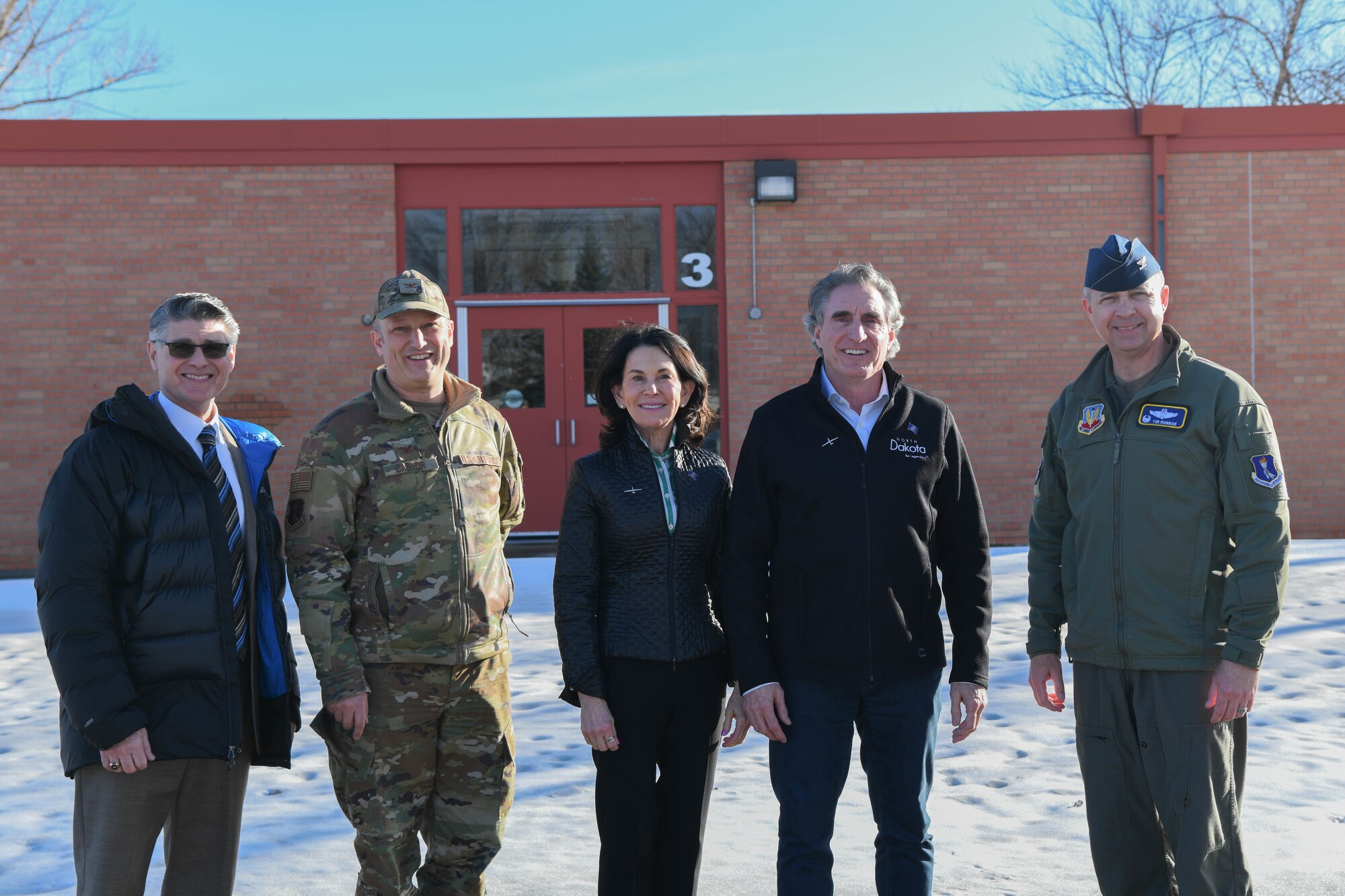 Two men in military uniforms pose for a photo with three others in front of a school.