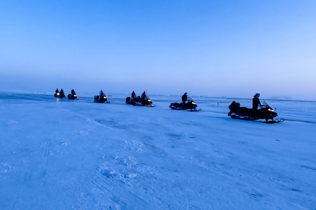 Soldiers drive a line of snowmobiles across the ice in an open area against a blue sky.