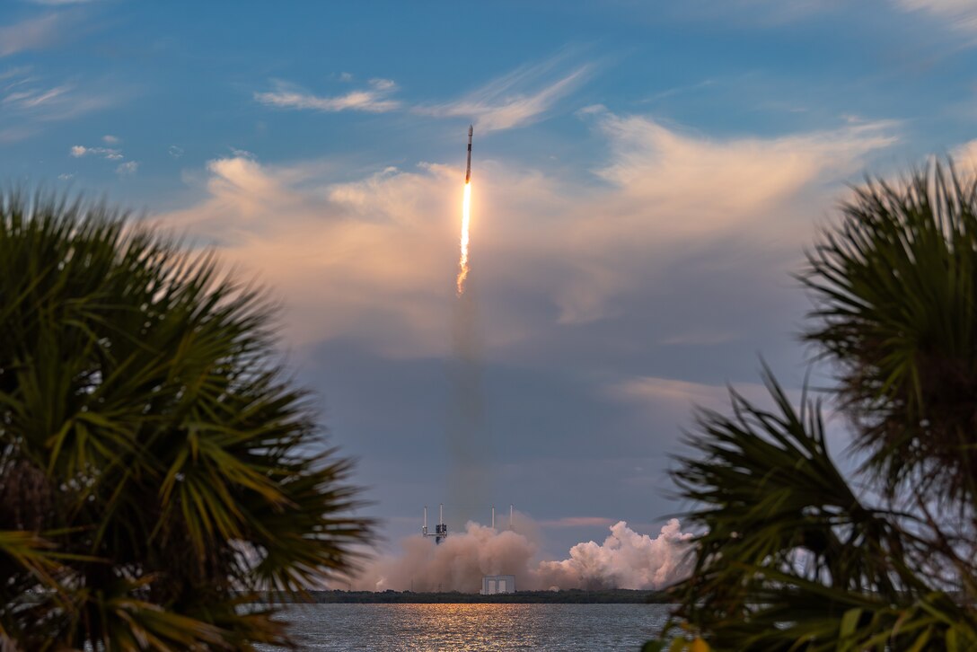 A rocket launches leaving a trail of smoke and fire against a blue sky with wispy clouds. Palm trees and water can be seen in the foreground.