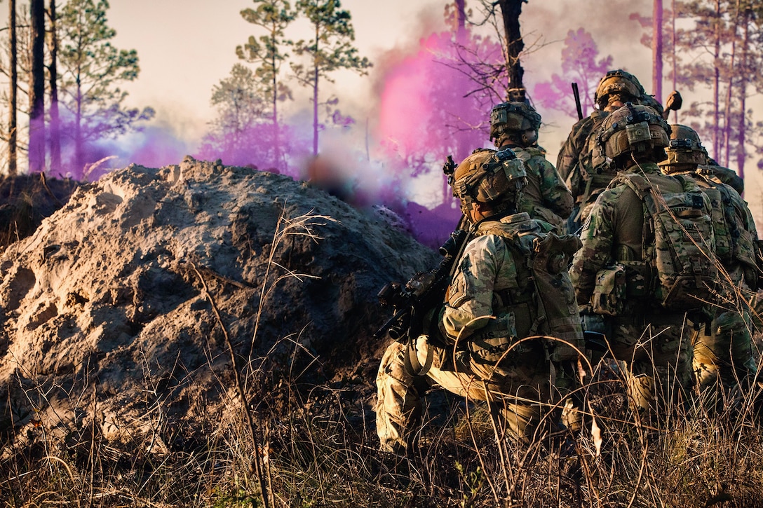 Soldiers with take position behind a small mound of dirt in a tree-lined area during training as purple smoke arises in the distance.