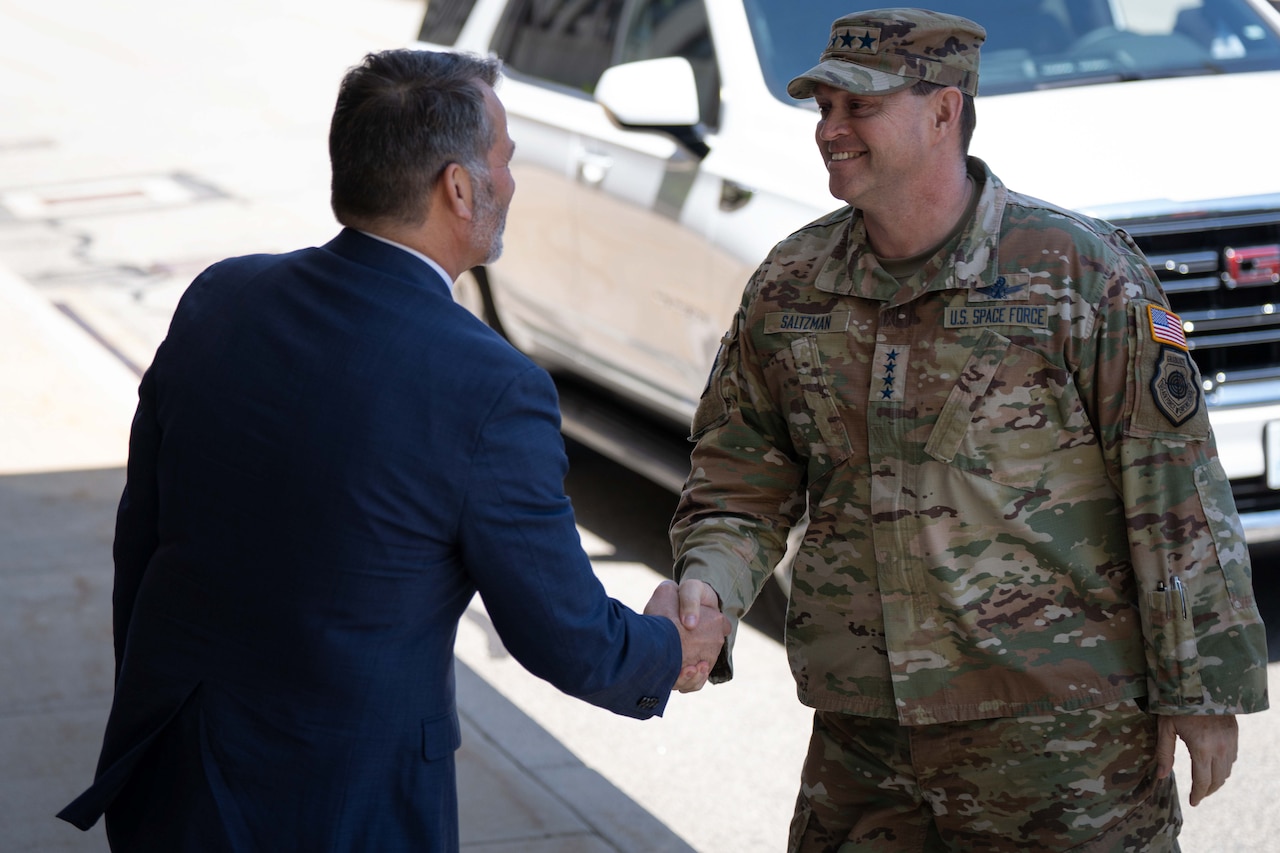 A man in a camouflage military uniform shakes hands with a man in a blue business suit.
