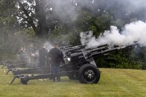 Army soldiers in dark ceremonial uniforms are firing black cannons that are placed on a green lawn.