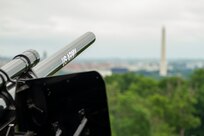 A black cannon with the words U.S. Army on the barrel is pointed out over an area with trees and the Washington Monument is seen in the distance.