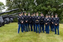 Army soldiers in dark ceremonial uniforms pose for picture in front of cannons that are placed on a green lawn.