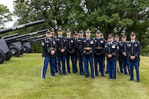 Army soldiers in dark ceremonial uniforms pose for picture in front of cannons that are placed on a green lawn.