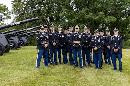 Army soldiers in dark ceremonial uniforms pose for picture in front of cannons that are placed on a green lawn.