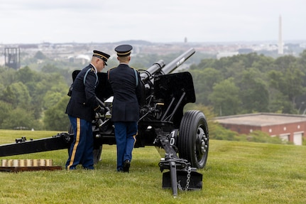 Army soldiers in dark ceremonial uniforms are firing black cannons that are placed on a green lawn that overlooks the white marble Washington Monument.