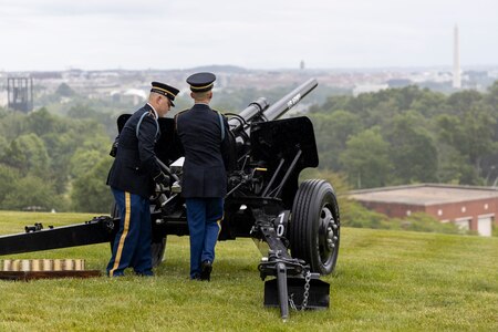 Army soldiers in dark ceremonial uniforms are firing black cannons that are placed on a green lawn that overlooks the white marble Washington Monument.